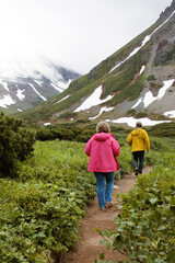Senior couple in bright tourist jackets is hiking in lush greenery near spectacular mountains with snow. Trekking in wild places. Happy active retirement concept. Old people healthy habits.
