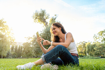 Young woman in casual clothing sitting on the grass in a city park and using her smartphone.