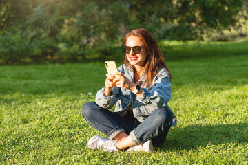 Fototapeta premium Young woman in casual clothing sitting on the grass in a city park and using her smartphone.