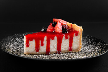 New York cheesecake with strawberries and syrup on a dark plate on a black background