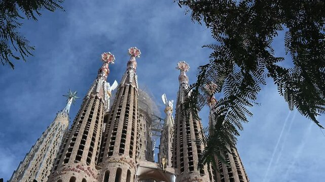 The camera pans up the Sagrada Familia, revealing its towering spires, intricate facades, sculpted details, arched entrances, and ongoing construction.