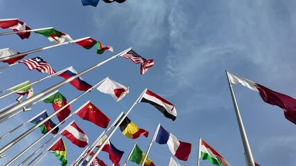 A collection of international flags waving on tall poles against a bright blue sky, symbolizing global unity and diversity.