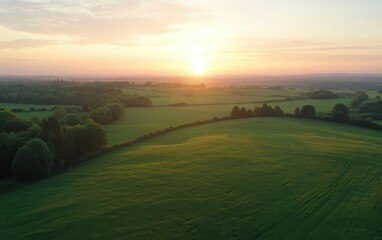Obraz premium Sunrise Over Verdant Farmland: A wide aerial view of the UK countryside, where fields of various shades of green stretch out beneath the rising sun. 