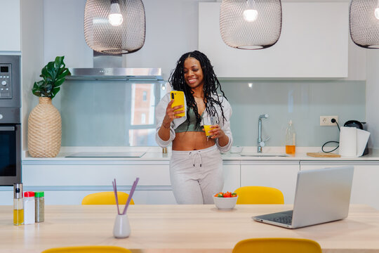 Young woman stands in her modern kitchen, enjoying a healthy breakfast while using her smartphone