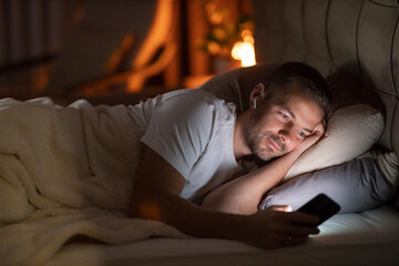 A man is lying in dark bedroom at night and watching podcast on cellphone during bedtime.