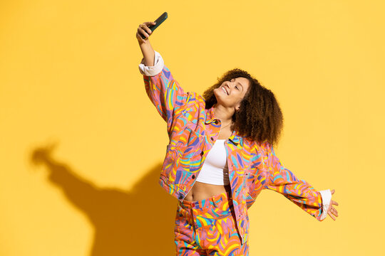 Happy young woman taking selfie through smart phone against yellow background
