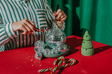 Woman cutting Christmas balls kept in plate on red table near candy cane