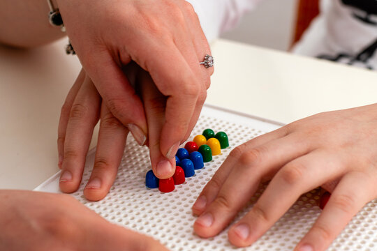 A girl engages in educational play with her teacher, who helps her arrange colorful pegs, focusing on finger movements and coordination.