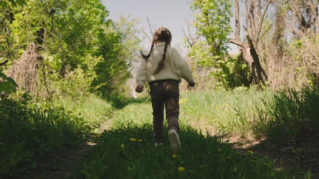 A girl is walking through a forest. The girl is wearing a white jacket and brown pants. The forest is lush and green, with trees and grass. The girl appears to be enjoying her walk in the woods
