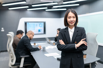 Businesswoman standing poses, group meeting around a table in modern office, documents, laptops and graphs are placed, everyone brainstorming, planning sales, digital marketing and growth strategies.