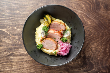 A beautifully plated dish featuring tender slices of pork, creamy mashed potatoes, vibrant pink pickled onions, and fresh herbs, served in a black bowl against a warm wooden background.