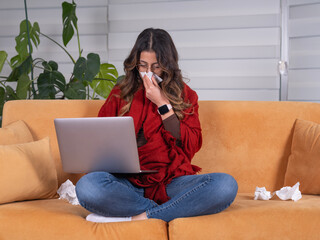 Obraz premium Sick woman using laptop, full body view caucasian sick woman using laptop sneezing blowing her nose to napkin sitting at sofa in home freezing warming wrapped with plaid. Flu concept idea.