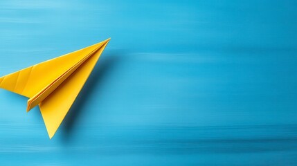 A bright yellow origami airplane sits on a light blue wooden surface. The airplane is positioned in the center left of the image, against a backdrop