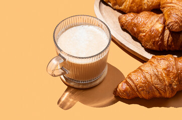 Cappuccino or latte coffee in a cup on an orange background with fresh croissants on a wooden plate.