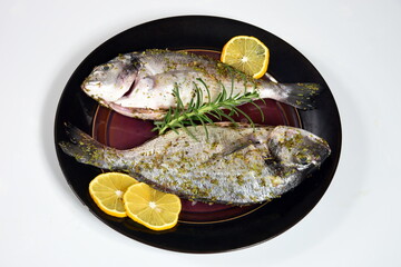 Two unbaked fresh dorada in a dish, decorated with lemon and spices. Dorado recipes, white background