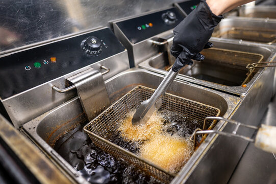 A chef uses tongs to lift a basket of fried food from a commercial deep fryer. The kitchen setting features multiple fryers and bubbling oil, emphasizing a busy culinary environment.