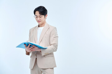 A young male teacher wearing a suit and glasses is holding a notebook in a classroom and conducting...