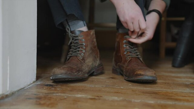 A man is putting on his brown boots. The boots are old and worn, but they look comfortable. The man is sitting on a chair while he puts on his boots