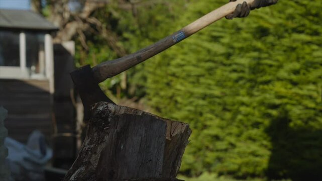 A man is holding a log in his hand. The log is on top of a stump. The stump is in front of a house. The man is wearing a black glove