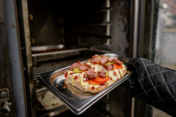 A close-up of a hand wearing an oven mitt, holding a tray of freshly cooked pizza with pepperoni and pickles, emerging from an oven with racks in the background. Perfect for food-related concepts.