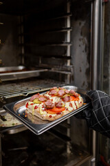 A close-up of a hand wearing an oven mitt, holding a tray of freshly cooked pizza with pepperoni and pickles, emerging from an oven with racks in the background. Perfect for food-related concepts.