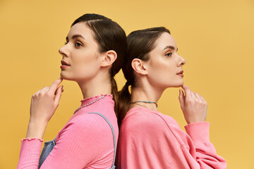 Beautiful twins pose back to back in stylish outfits against a vibrant yellow background