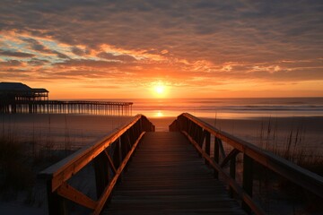 Fototapeta premium Sunrise illuminating wooden walkway leading to the beach with pier under colorful sky at dawn
