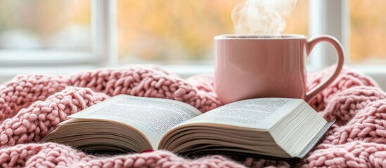 Close-up of an open book and a steaming coffee mug on top, with a pastel pink knitted blanket by the window.