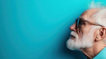 Fototapeta premium An elderly man with a white beard and glasses gazes thoughtfully to the side, framed by a smooth blue backdrop that emphasizes his reflective expression and wisdom.