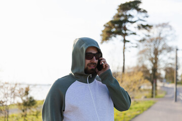 Handsome bearded man in sunglasses and a hood walking along a quiet seaside pedestrian road, talking on his mobile phone, exuding a stylish, modern, and relaxed urban lifestyle by the coast