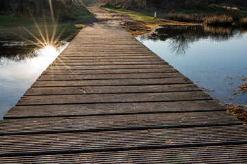 A rustic wooden footbridge stretches over a calm pond on Schiermonnikoog, reflecting the warm glow of the setting sun.