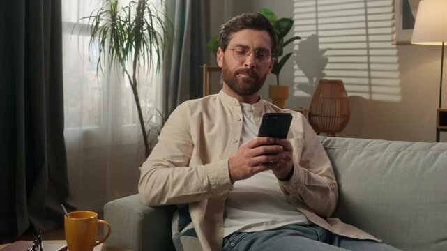 Man thoughtful dreaming Caucasian male homeowner tenant freelancer at home using mobile phone app online shopping in apartment smiling guy think on couch sofa relax browsing smartphone gadget addict
