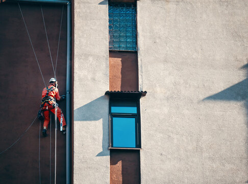 Industrial climber in red uniform on ropes fixing pipes on urban building facade. Professional safety equipment and geometric textures.