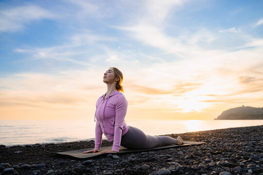 Woman practicing yoga cobra pose on a stone beach at sunset or sunrise. Exercise, fitness, stretching, workout, pilates, wellness outdoors.