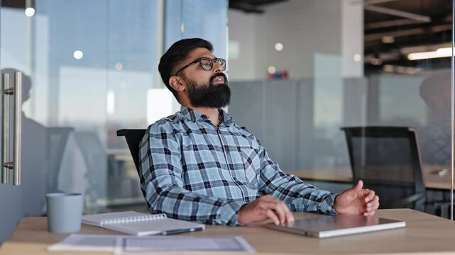 Bearded businessman closes laptop, leans back in office chair, puts hands behind head and rests in modern office.