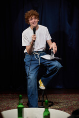 Young man sitting on stool while engaging audience with microphone in hand, smiling and holding...