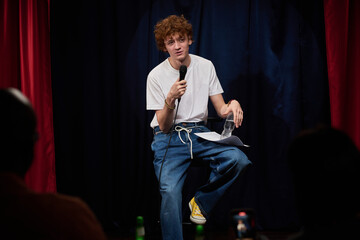 Person performing stand-up comedy with microphone in hand while holding water bottle in other hand on stage with red curtains and audience in foreground