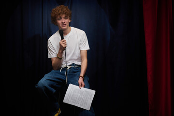Young man standing on stage with paper and microphone performing comedy routine with a focused...