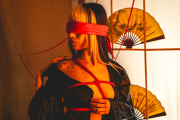 Sexy and beautiful young woman with blonde hair, black kimono and panties, and red shibari ties and ropes, red blindfold, holding a paper fan in a warm japanese room