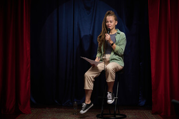 Young woman performing stand-up comedy on stage under spotlight while holding microphone and...