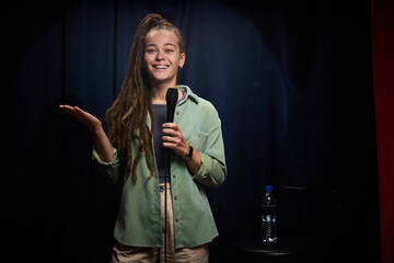 Woman with long hair, wearing casual outfit, standing with microphone on stage and smiling while...
