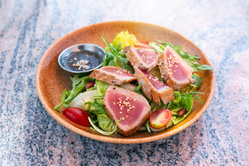Seared sliced meat ​​fish with mixed green fresh vegetable. Tuna Loin salad with honey orange sauce with white sesame seeds in brown ceramic plates on marble table.