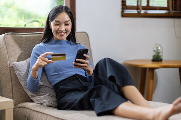 Young Asian woman at her happy home is shopping online on a smartphone and paying with a credit card via mobile payment.