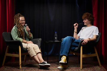 Two people sitting on chairs while recording a podcast with microphones in hand, appearing to share...