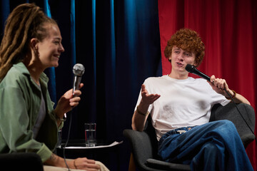 Two individuals engaging in an interview conversation, each holding microphones and seated on stage...