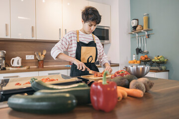 Young girl wearing apron, slicing tomatoes on cutting board in modern kitchen, surrounded by fresh...