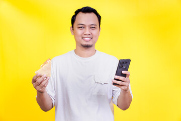 A cheerful young asian man holds food in one hand and a smartphone in the other against a yellow background. international foods day concept