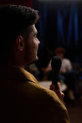 Man standing on stage holding microphone in hand, seen from behind. Blurry audience visible in...