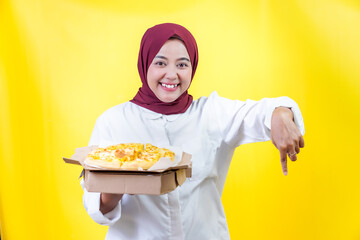 A cheerful asian woman in a hijab happily presents a delicious pizza against a bright yellow background