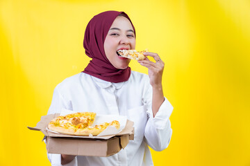 An asian woman enjoys a slice of pizza while holding a box of pizza in front of a bright yellow...